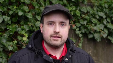 A man standing in front of a brown fence with foliage growing over it smiles at the camera. He is wearing a black cap with a black coat. He has a red polo top on underneath. 