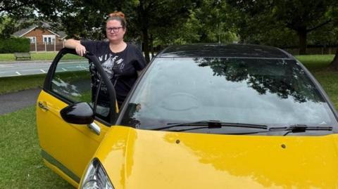 Emma Drinkwater with her brown hair tied up in a ponytail and wearing glasses with a serious expression. She has a black top on with a white pattern. She is leaning against her yellow car with the driver's door open. 