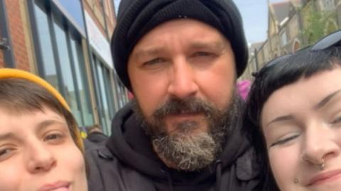 A man with a grey beard and moustache wearing a black beanie hat and black hoodie. A young woman is stood on either side of him. One has short brown hair and a yellow beanie hat, the other has black hair with sunglasses on her head and face peircings.