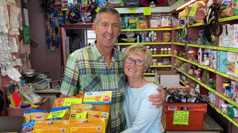 Terry has short hair and is wearing a green checked shirt. He is standing with his arm around Orla who has short blonde hair and is wearing glasses and a blue and white striped top. They are both stood at the counter in the pet shop smiling at the camera. 