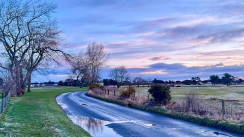 A country scene in Nairn in the highlands with fields and a country road winding away in to the distance. There is a pale sky with pinks and greys. No people or buildings are visible
