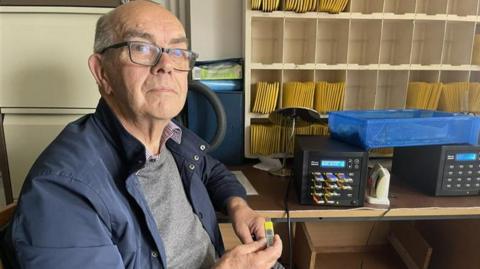 A man wearing glasses, a blue jacket and a grey sweater looks towards the camera. He is holding a yellow USB stick. Behind him is a set of shelves that is partially filled with yellow documents. In front of the shelving is a desk that has two black bits of electronic equipment on it.