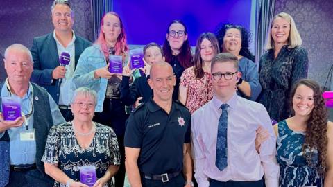 A picture of the winners from last years make a difference awards. There is a number of people holding purple trophies