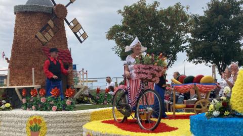 A series of floats used in the Battle of Flowers earlier this year. One has a windmill and someone dressed in a black cap, and red shirt. The other has someone dressed in a white hat, top, and red and a red and white striped dress, standing beside a bicycle full of flowers.
