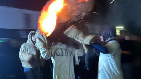 A man with a barrel on his back. It has flames coming from it and there are three other men crowded round. It is dark.