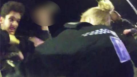 A still image from body-worn police camera footage, showing a female police officer with blonde hair and her black police-issue jacket, who appears to be wrestling with Declan Diedrick, who has thick hair and is gritting his teeth menacingly.