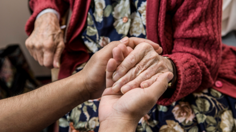 Two pairs of hands holding each other with the older pair of hands belonging to a woman with a red cardigan and a patterned dress