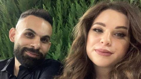 Ashraf and Julia both smiling for the camera, with a background of greenery. Ashraf has dark hair and a beard and is wearing a dark shirt, and Julia has lighter brown hair - long and wavy - and has brightly painted lips.
