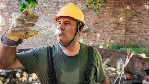 Man wearing a yellow safety helmet and sweaty green T-shirt drinking water from a plastic bottle