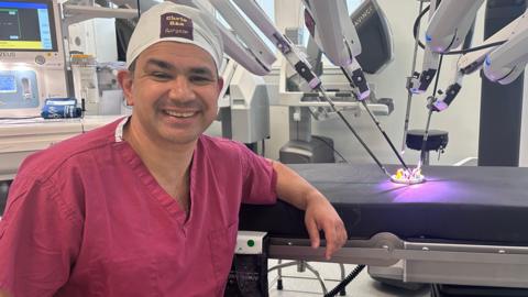 Chris Rao, colorectal consultant and surgeon at the Cumberland Infirmary in Carlisle. Rao is sat in front of a hospital bed with the robot arms simulating an operation on a prop. Rao is wearing a white fabric scrub cap with his name stitched on it in yellow and purple. He is wearing burgundy scrubs and is smiling at the camera. The robot has four arms with metal long and thin rods manoeuvring the prop.