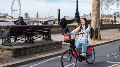Woman rides a Santander bike on a London road with Big Ben and the London Eye in the background. She smiles and wears a white coat and jeans. 