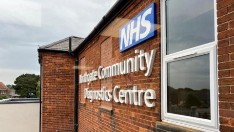 A view down the side of a brick building, with writing on the wall that says "Northgate Community Diagnostics Centre", beneath a blue and white NHS logo. A white plastic window is in the foreground on the right.