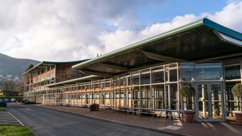 A modern glass-fronted office building with sweeping green roofs sits beside a quiet road. Behind it, rolling hills rise under a cloudy sky.