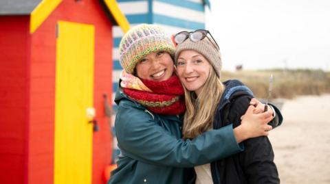 Two people stood on a beach in the winter. The woman on the right is wearing a big green coat and wooly hat. The woman on the right had a grey wooly hat and a dark coat. They are hugging, stood in front of a beach hut.