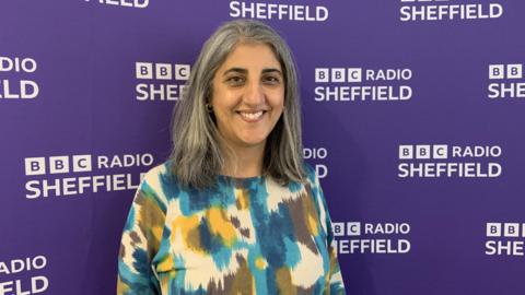 A woman with shoulder length grey hair is wearing a blue, yellow and white patterned top. She is standing in front of a purple wall with white writing saying BBC Radio Sheffield
