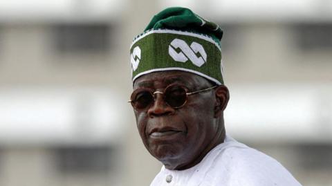A head and shoulders image of Bola Tinubu wearing dark, round glasses and a green cap. 