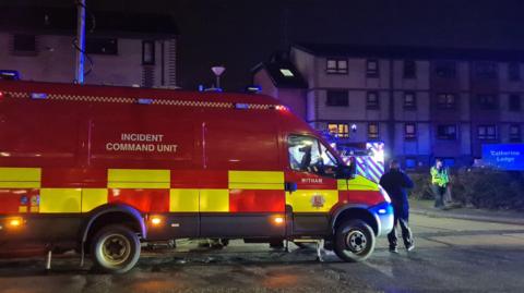 A red and yellow incident command unit van is parked outside a residential area at night-time. To the right of the vehicle is a police office and a sign reading 'Catherine Lodge'.