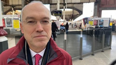 Jonathan Denby, with close cropped greying hair, standing near the glass barriers at Norwich station. He is wearing a red jacket, a white shirt with red tie, and wearing glasses. In the distance are destination boards, station platforms and a train.