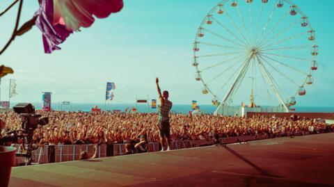 A performer stands on a stage facing a large crowd at an outdoor festival by the sea, with many people raising their hands. In the background, a large Ferris wheel rises above the crowd under a bright blue sky, while colourful flags and decorations add to the lively atmosphere.