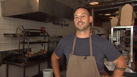 A man with brown hair and is wearing a navy t-shirt under a brown apron. He is in a kitchen with a wood fired pizza oven in the background
