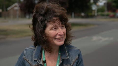 A woman with medium-length curly hair wearing a denim jacket and a green lanyard stands outdoors on a quiet street with trees and houses blurred in the background.