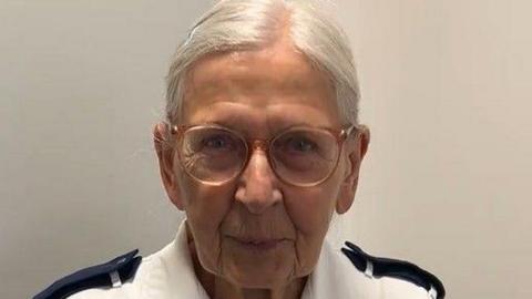 Iva smiles directly into the camera.  Her eyes are twinkling behind her clear light pink glasses and she wears a white nurses uniform.