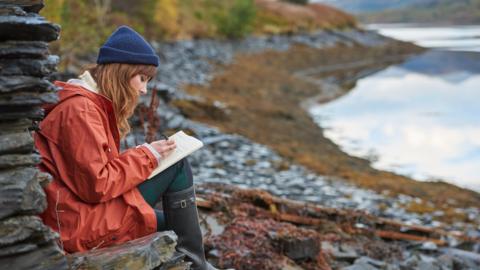 A woman in a red anorak, dark leggings, wellington boots and a blue beanie hat sits beside a river, writing in a journal.