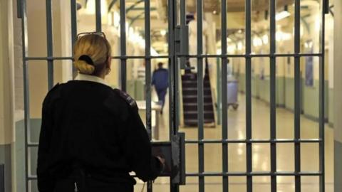 A blonde female prison guard in a black uniform has her back to the camera, she is unlocking a prison door