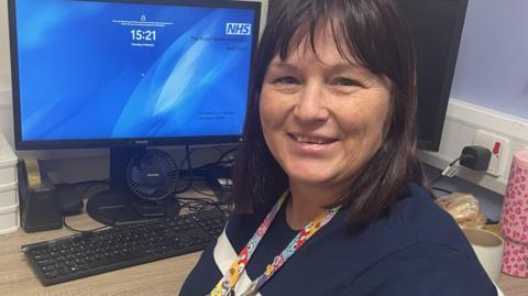 A woman with shoulder length dark hair and wearing a blue t- shirt sits next to a computer at work.  She has a lanyard round her neck which has teddy bear pictures on it. 