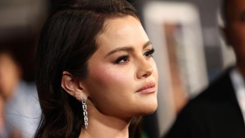 Selena Gomez at the premiere of Emilia Pérez. Selena is pictured from her right-hand side, her dark hair worn loose. She has brown eyes and wears a black dress with silver earrings. In the background are other people on the red carpet. 