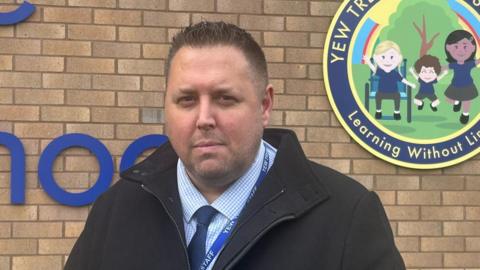 Jamie Barry is stood in front of the Yew Tree Primary School sign and brick building. He is wearing a chequered shirt and blue tie with a black coat on.