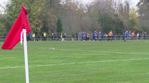 A scene from football being played at Faraday Road in Newbury, where Newbury FC (playing in orange and black) are playing against a team in a blue kit. A red corner flag is pictured on the left side of the picture, with a handful of spectators lining the pitch