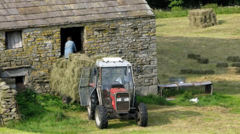 A stone barn on a rural farm with a red tractor parked outside, its front loader carrying a large pile of hay.