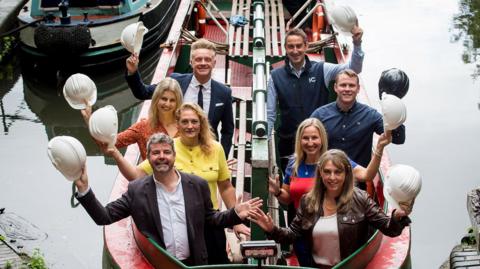 Eight people sit in a canal boat and hold up their own white builder's hat.