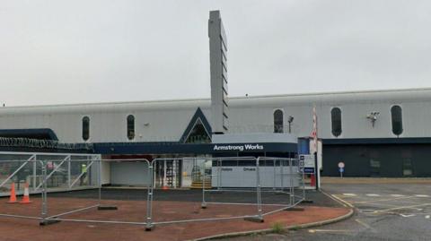 The Armstrong Works site on Newcastle's Scotswood Road. A gatehouse is located in front of the building which is made from grey corrugated metal.