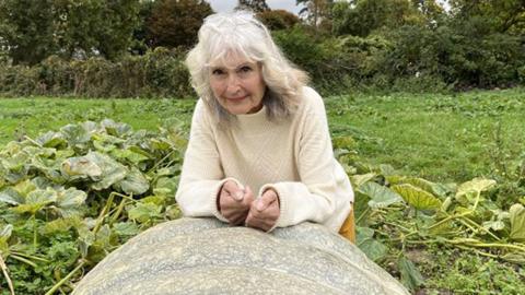 Frances Crickmore leaning on a very large light green pumpkin. She has white shoulder-length hair and is wearing a cream Arran style jumper. Behind her are pumpkin vines, grass and beyond the a hedge. 