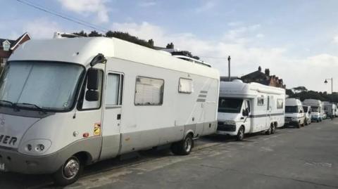 A row of light coloured campervans, parked up bumper to bumper on a road. Above can be glimpsed roofs of buildings. In front is a wide tarmacked road or pavement. 