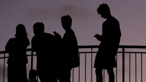 Silhouettes of young people on their phones standing against a barrier