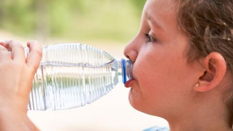 Close-up of the face of a girl standing in casual clothes drinking a bottle of water in a park, side view - stock photo