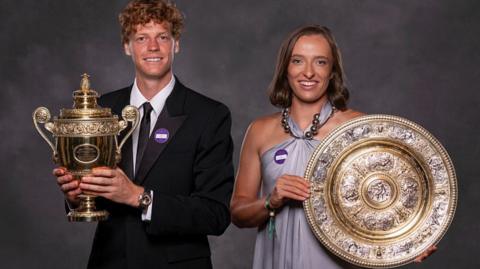 The Wimbledon singles champions pictured together. Jannik Sinner of Italy  (left) holds the Gentlemen's Singles Trophy — a stately gold cup with ornate handles and a pineapple-shaped finial on top, steeped in tradition since 1887. On the right,  Iga Swiatek of Poland, the Ladies' Singles champion presents the Venus Rosewater Dish, an intricately decorated silver-gilt salver