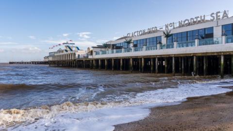 A side-angle view of Clacton Pier extending over choppy, brown ocean waves onto a sandy beach under a partly cloudy sky. The large building on the pier features large windows and a sign on top that reads 'CLACTON PIER - NO. 1 NORTH SEA,' with amusement park rides visible further down the deck