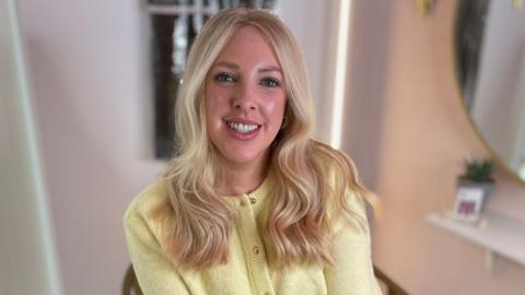 Laura Rush has long wavy blonde hair. She is smiling, sitting in a hairdressers chair and wearing a light yellow cardigan.