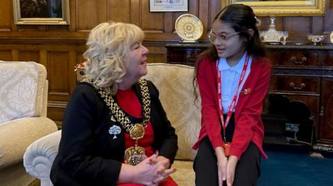 A blond-haired woman wearing a black jacket, red dress and gold mayoral chain sitting on an armchair talking to a young girl with dark hair and glass, wearing a red school cardigan, white polo shirt and black trousers perched on the arm of the chair. 