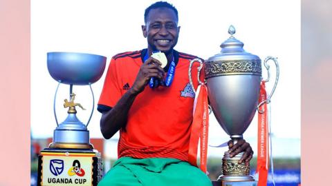 Abubabakar Lawal, in a red football jersey, holding a medal and sitting in between two trophies