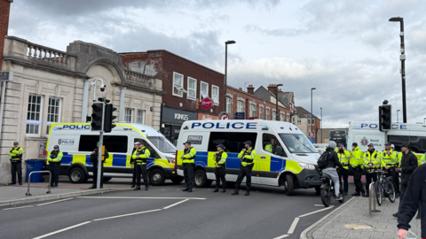 Two police vans and at least 10 police officers hi-vis gear parked across a road