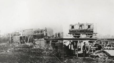 Old train carriages on a viaduct, some partially off the rails. Other carriages are destroyed underneath the viaduct in the black and white image.
