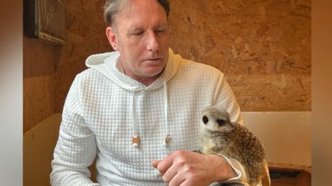Landlord Chris Collier pictured with a meerkat at the Royal Oak Inn and Meerkat Retreat in Brockham, Surrey. Chris is wearing a white fleece and has a meerkat on his arm. He is sitting in the enclosure.