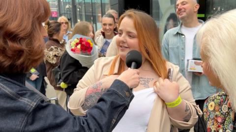 A woman with dyed ginger hair pulls her top to one side to show a tattoo below her collar bone 