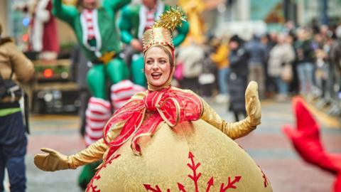 A woman in a Christmas pudding costume costume smiles as she walks down a street. 