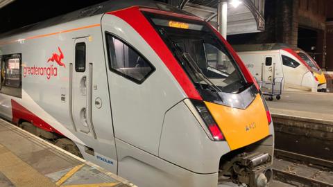 A white train with a red and yellow front. It has a red Greater Anglia signage on it and a red rabbit. The train is stationary on a train track by a platform.